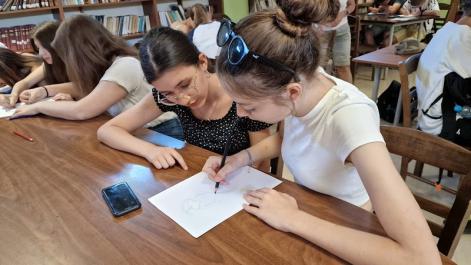 Młodzież w bibliotece siedzi przy stoliku, coś piszą, 