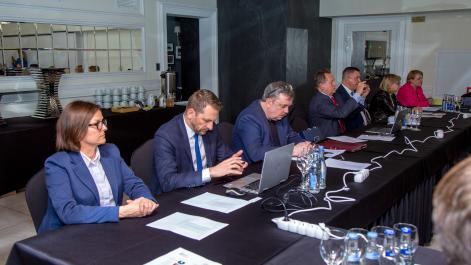 Członkowie posiedzenia siedzą za stołem, pracują na laptopach