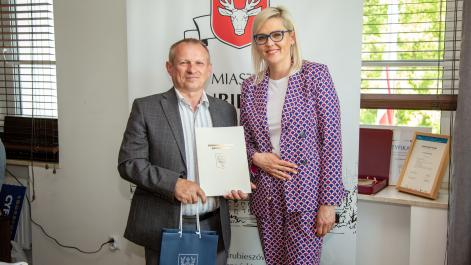 Marta Majewska pozuje do zdjęcia z trenerem sekcji podnoszenia ciężarów. Trener trzyma w ręku teczkę z herbem miasta i upominek w granatowej torbie, w tle widać baner Miasta Hrubieszów