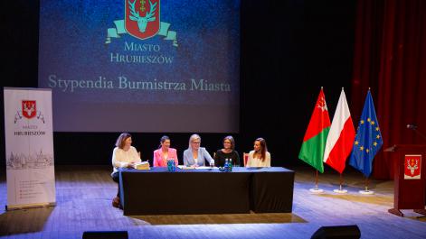 Cztery kobiety siedzą przy stole prezydialnym, obok stoją flagi, trwa uroczystość wręczenia stypendiów Burmistrza miasta