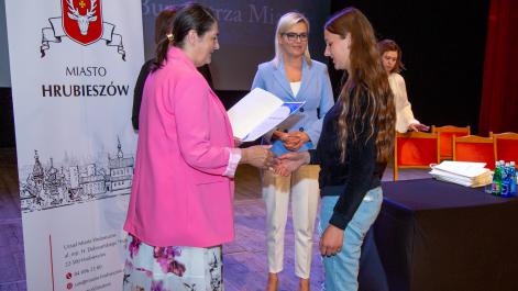 Kobieta w różowej marynarce wręcza dziewczynce dyplom, w tle widać drugą kobietę