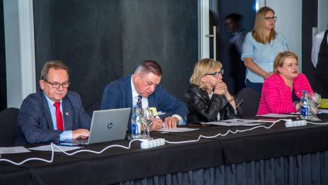 Kilka osób siedzi przy stole, jedna jest zasłuchana, jedna coś piesze, jedna pracuje na komputerze 