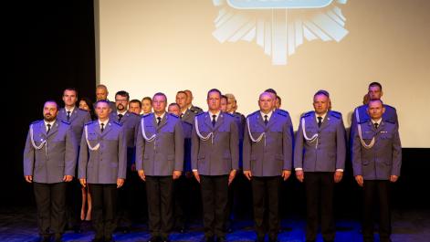 Policjanci stoją w rzędach, czekają na odznaczenia, w tle widać wyświetlone logo policji