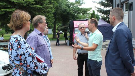 Mężczyzna tłumaczy coś pozostałym uczestnikom spotkania, wszyscy stoją na zewnątrz, w tle widać namioty promocyjne